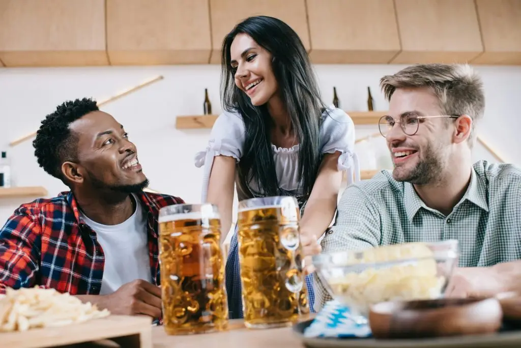 cheerful waitress in traditional german costume serving beer for multicultural friends in pub